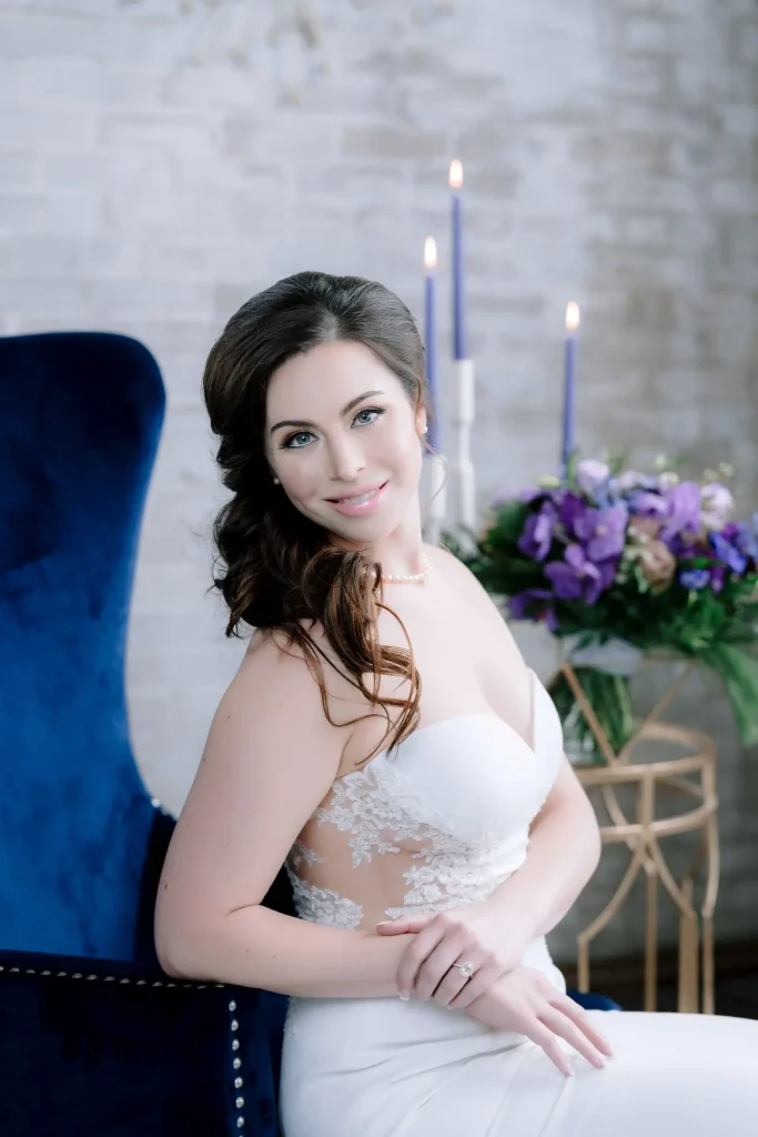 bridal portrait in studio with tight curls, glam makeup, blue chair and blue candles with flower bouquet in the background